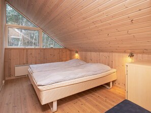 A bedroom with a wooden sloped ceiling, a large window, a bed, a bedside table, and a heater.