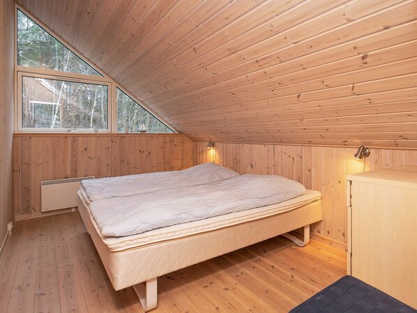 A bedroom with a wooden sloped ceiling, a bed, a heater, and a window with a view of trees.
