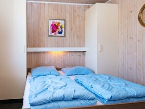 A single bed with a blue duvet and pillows, a wooden wall with a framed picture, and a white bedside table.