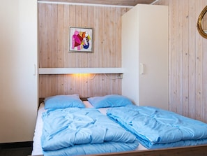A single bed with blue bedding, a wooden wall panel, a white shelf, and a framed picture above the bed.