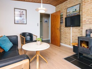 A cozy living room with a black leather sofa, a wooden chair, a round table with a plant, a brick wall, a TV, and a wood-burning stove.