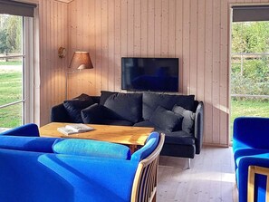 A room with a wooden paneled wall, a flat-screen TV mounted above a dark-colored sofa, a wooden coffee table with a magazine, and blue upholstered chairs. - 4 Star Holiday Home in Vaeggerlose (Vaeggerlose)