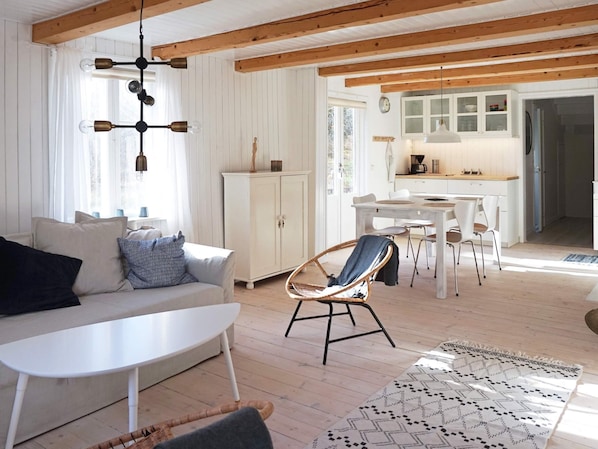 A modern living room with a white sofa, a small white table, a wicker chair, and a patterned rug. There is a kitchen area with white cabinets and a dining table. - 6 Person Holiday Home in Hallabro (Hallaröd)
