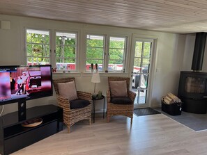 A cozy living room with two wicker chairs, a TV, a fireplace, and large windows with a view of greenery.