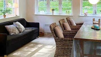 Una habitación con sofá, sillas de mimbre, una mesa y ventanas que dan a zonas verdes.