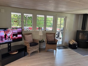 Een gezellige woonkamer met twee rieten stoelen, een televisie, een open haard en grote ramen met uitzicht op groen.