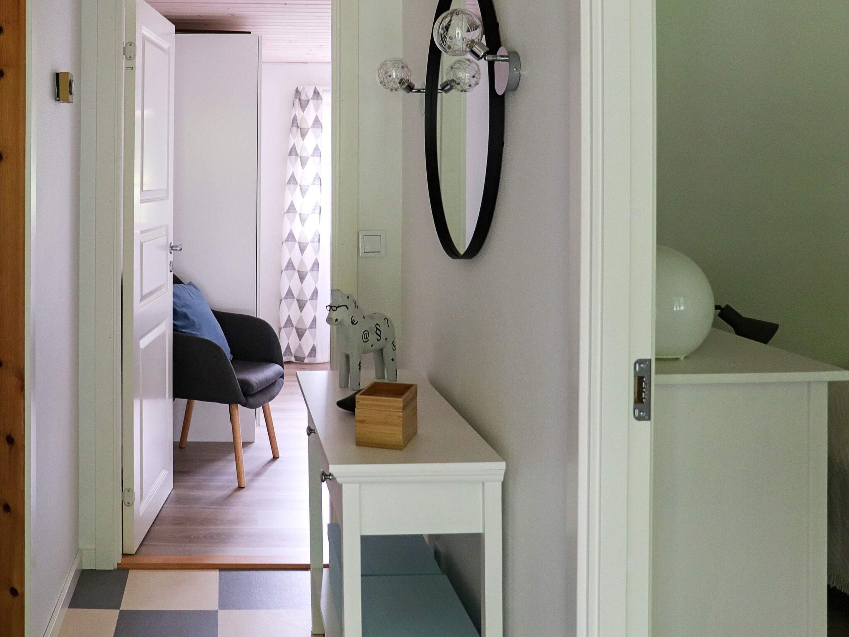 A room with a bed, a chair, a small table with a box, a wall-mounted light fixture, and a door leading to another room.