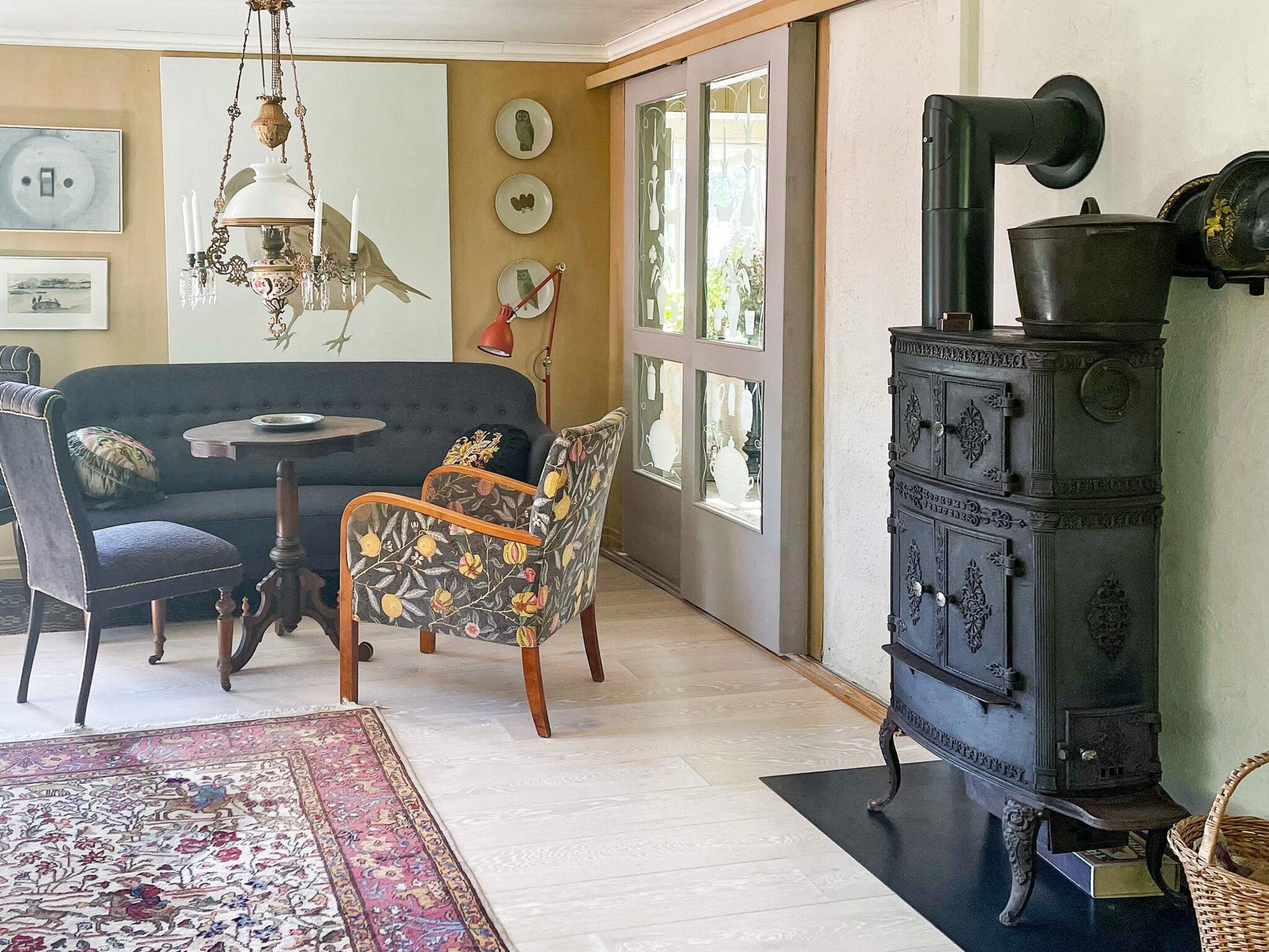 A room with a sofa, a patterned armchair, a chandelier, a vintage stove, and a large window with decorative plates.