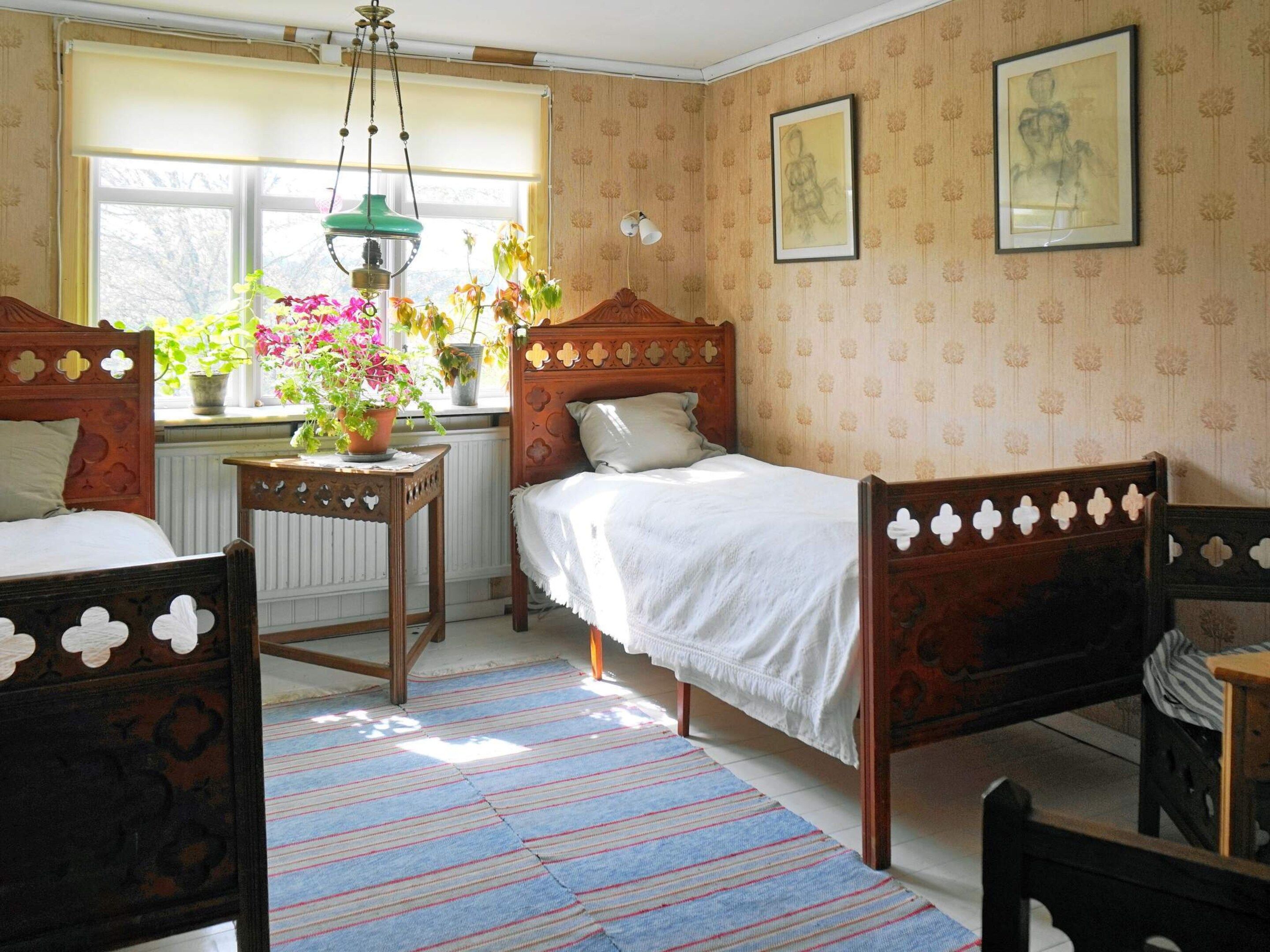 A room with two wooden beds, a window with plants, a radiator, and patterned wallpaper.