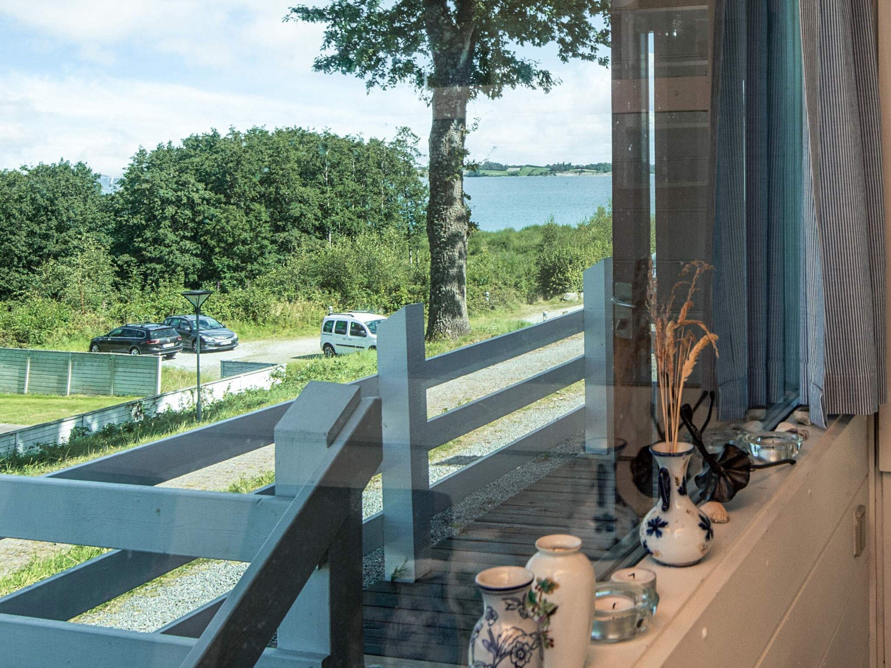 Vista desde una ventana sobre un lago, una terraza de madera y un estacionamiento con coches.