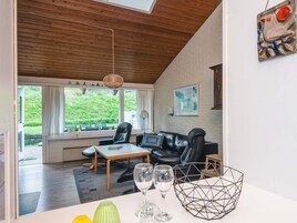 A modern living room with a wooden ceiling, a black leather sofa, a wooden coffee table, and a view of a green landscape through large windows.
