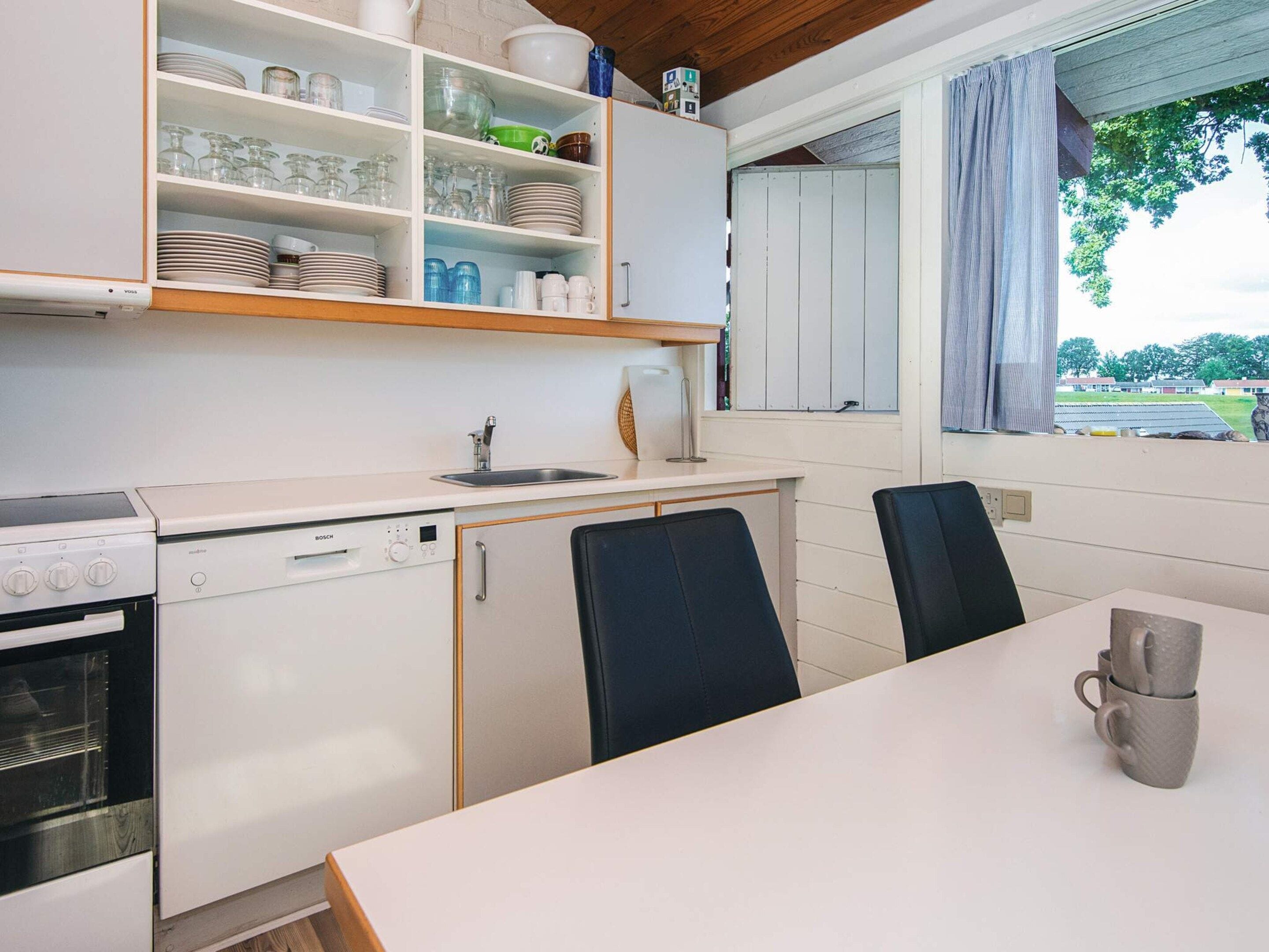 Cozinha compacta com armários brancos, máquina de lavar louça Bosch, fogão e área de refeições com duas cadeiras e uma mesa.