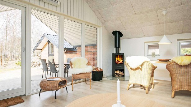 A spacious living room with a large glass door leading to an outdoor area, a black wood-burning stove, wicker furniture, and a round wooden table. - 4 Person Holiday Home in Laeso (Læsø)