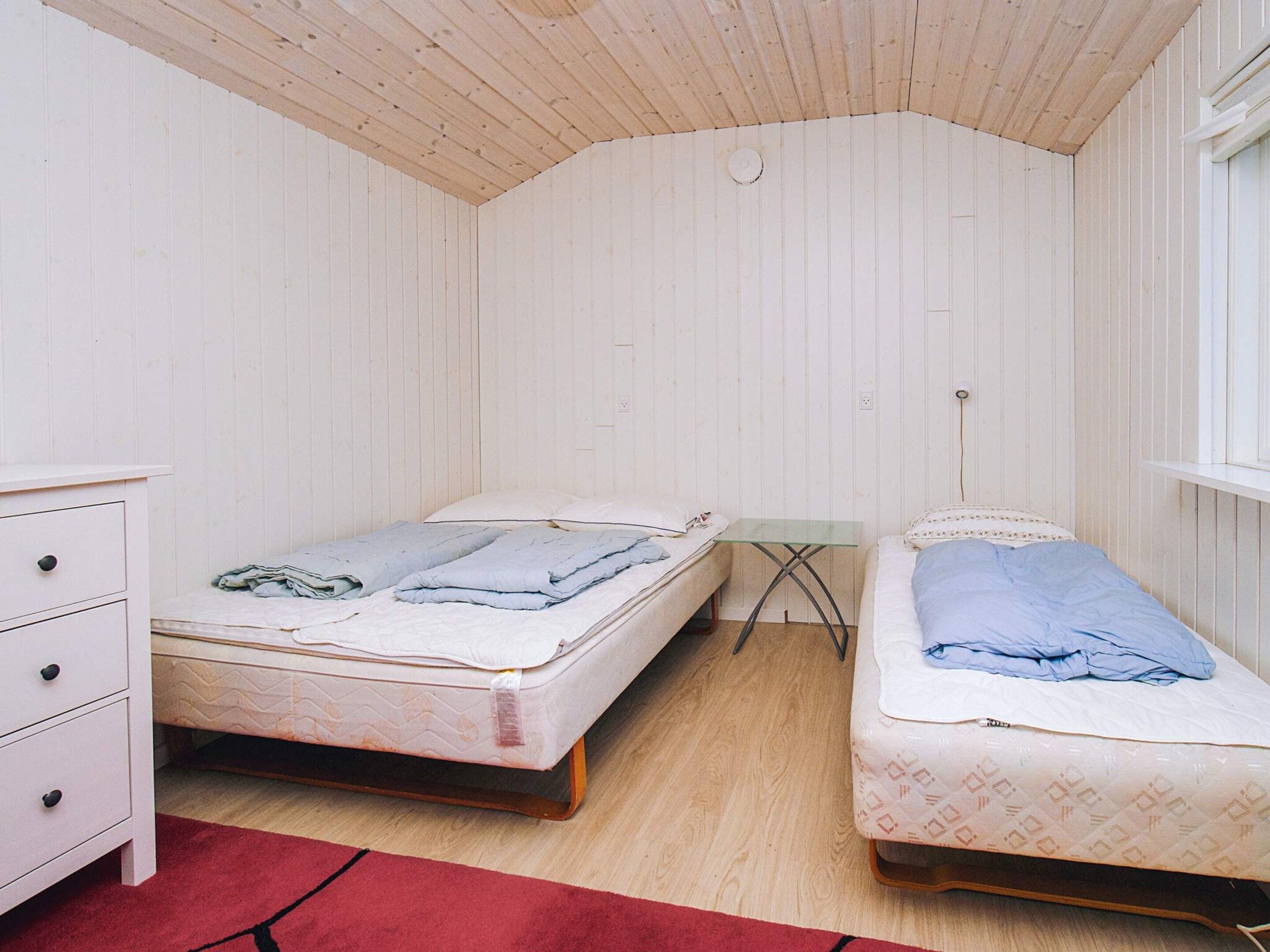 A room with two beds, a small table, a dresser, and a red rug.
