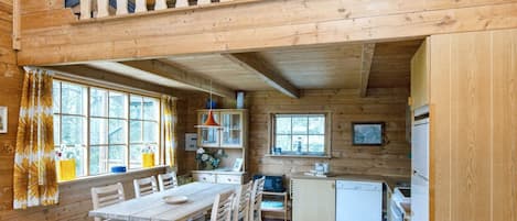 A wooden cabin interior with a dining area, kitchen, and a loft space with a balcony.