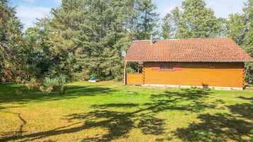 A single-story wooden cabin with a tiled roof, surrounded by a grassy yard and dense trees.