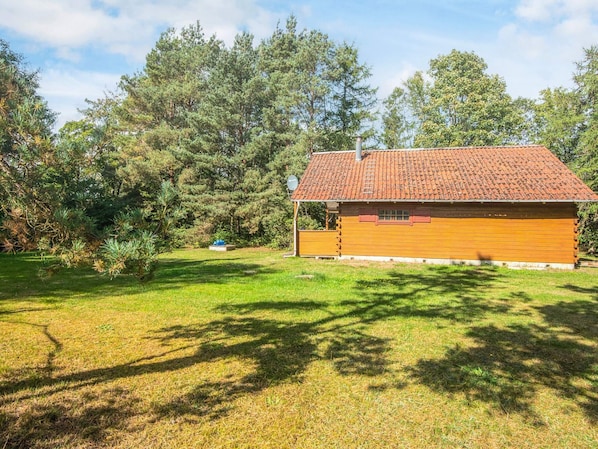 Une petite cabane en bois à un seul étage, avec un toit en tuiles, entourée d’une cour verdoyante et d’arbres denses.
