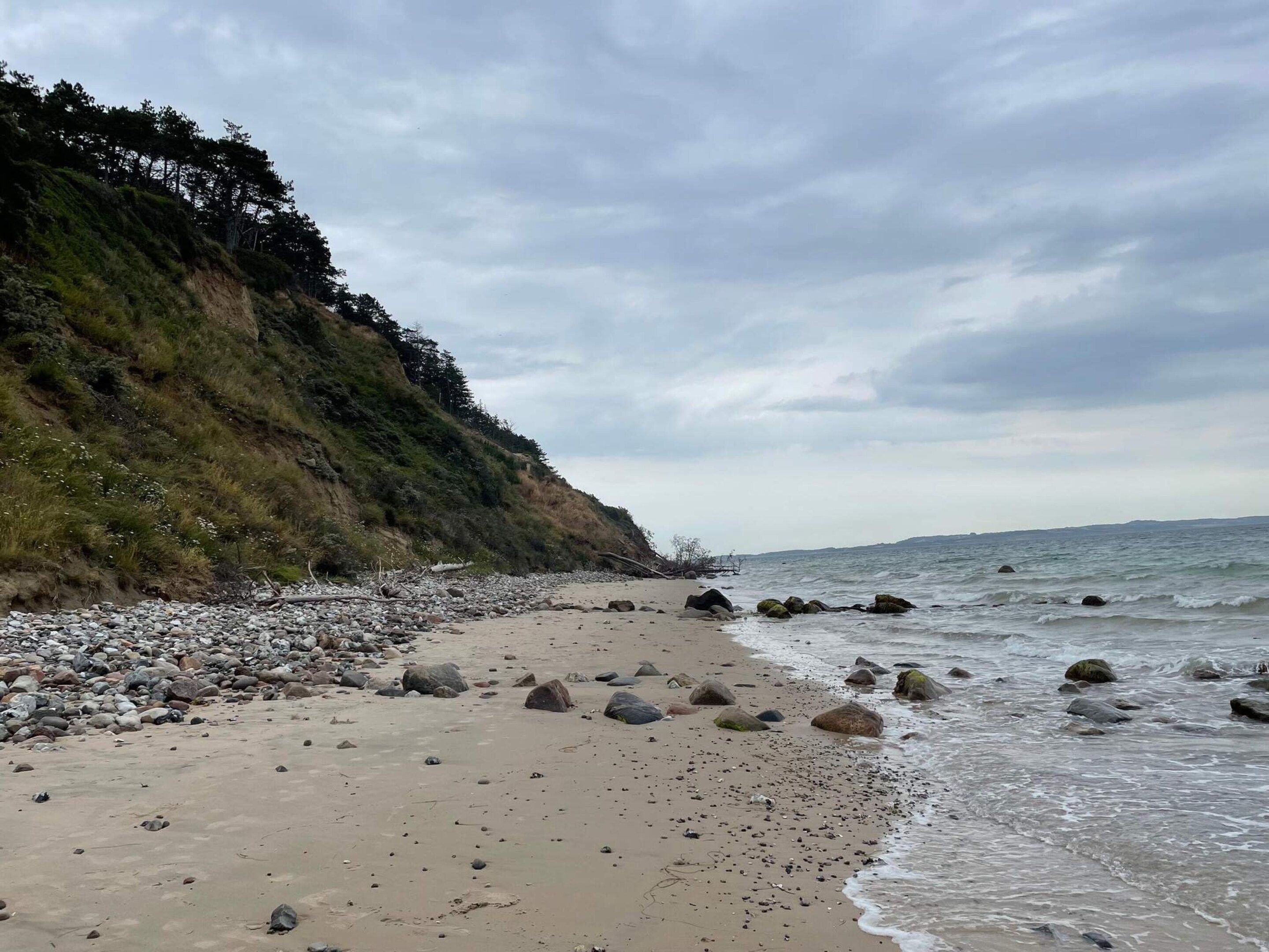 A beach with pebbles and rocks, a grassy cliff, and the ocean.