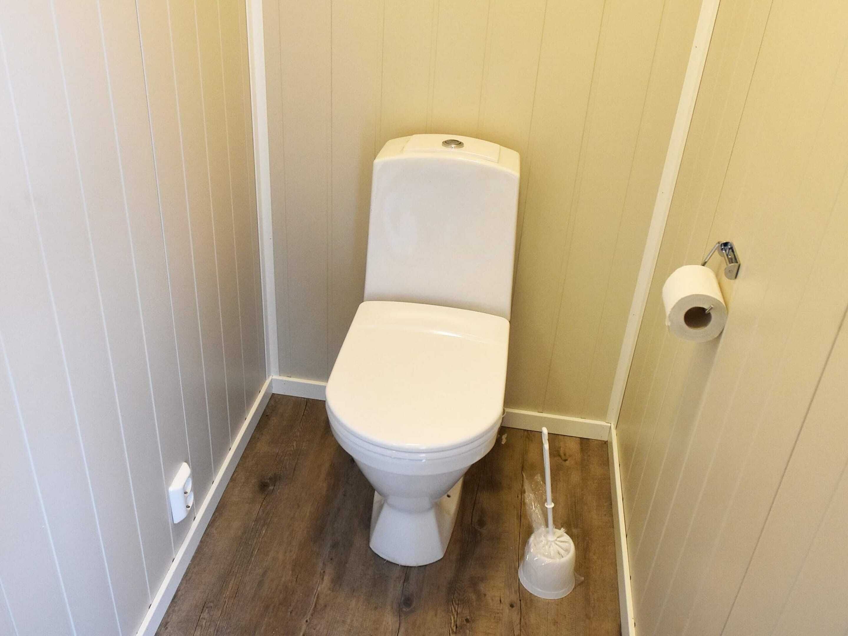 A small, white toilet in a corner with a wooden floor and a toilet paper holder on the wall.