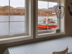 Une pièce dotée d’une grande fenêtre donnant sur un plan d’eau, avec un bateau rouge amarré à l’extérieur, et une table blanche recouverte d’une nappe à motifs.