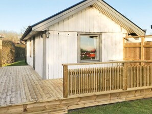 A wooden cabin with a deck, a window with a view of flowers, and a fenced area.