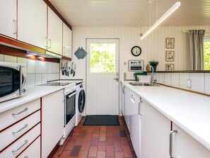 Une cuisine compacte avec des meubles blancs, un four à micro-ondes, une cuisinière et un lave-linge. Il y a une horloge au mur et une affiche indiquant le mot de passe du Wi-Fi.