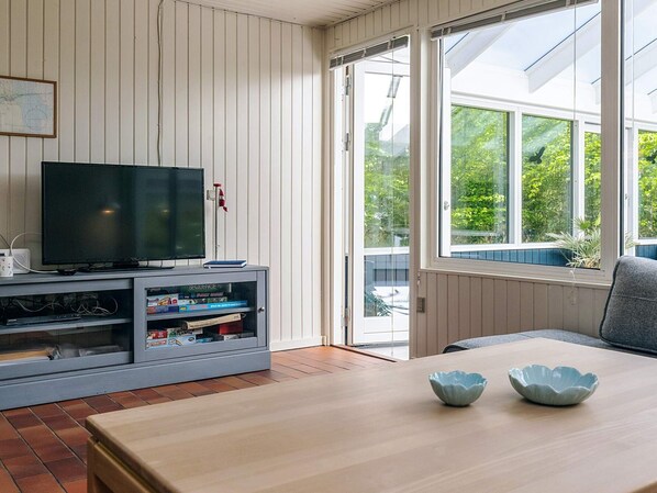 Un salon avec un téléviseur posé sur un meuble, une vitrine contenant des livres, une table à manger avec deux bols, et une porte-fenêtre coulissante donnant sur l’extérieur.