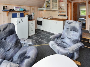 A compact kitchen with a refrigerator, stove, and sink. There are two upholstered chairs and a small table.