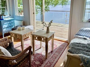 A room with a view of a lake, featuring a wicker chair, a small table with a vase, and a sofa with a blanket.