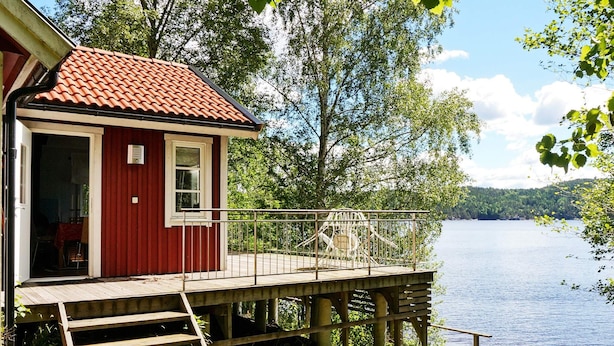 A red cabin with a wooden deck overlooking a lake, a metal railing, and a set of stairs leading to the entrance. - 4 Person Holiday Home in Bullaren (Bullaren)