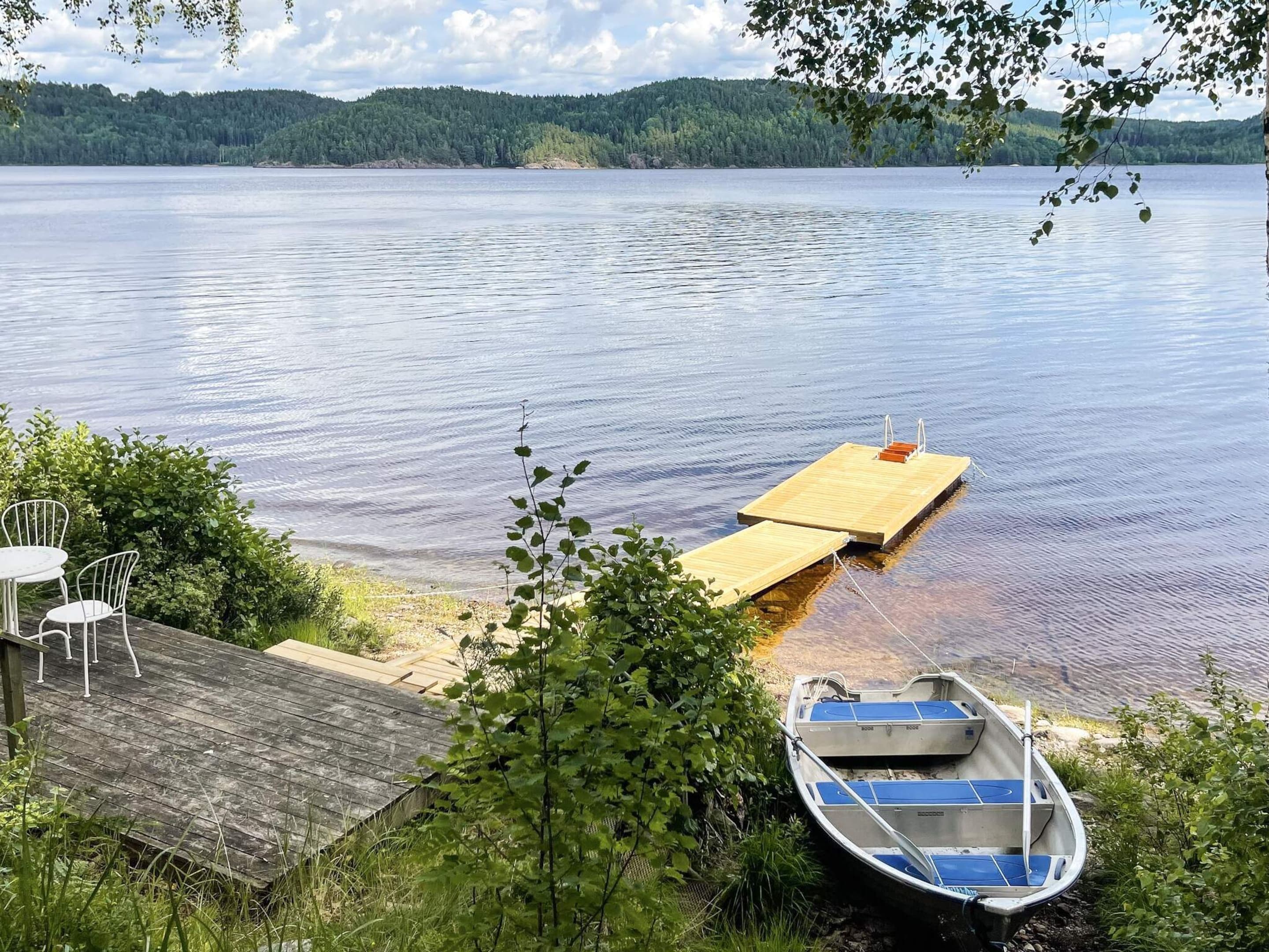 A lakeside dock with a small boat, a wooden deck, and a table with chairs.