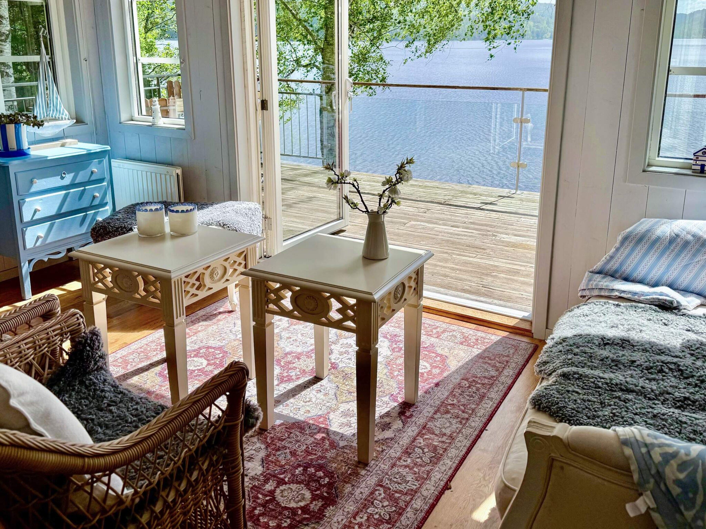 A room with a view of a lake, featuring a blue dresser, two white tables, a wicker chair, and a bed with a blue and white striped blanket.