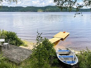 Une jetée en bois s’étendant sur un lac paisible, un petit bateau amarré à son extrémité, et une table avec des chaises sur la jetée.