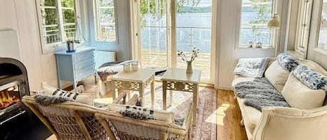 Una sala de estar acogedora con chimenea, muebles de mimbre, un sofá blanco y grandes ventanales con vista a un lago.