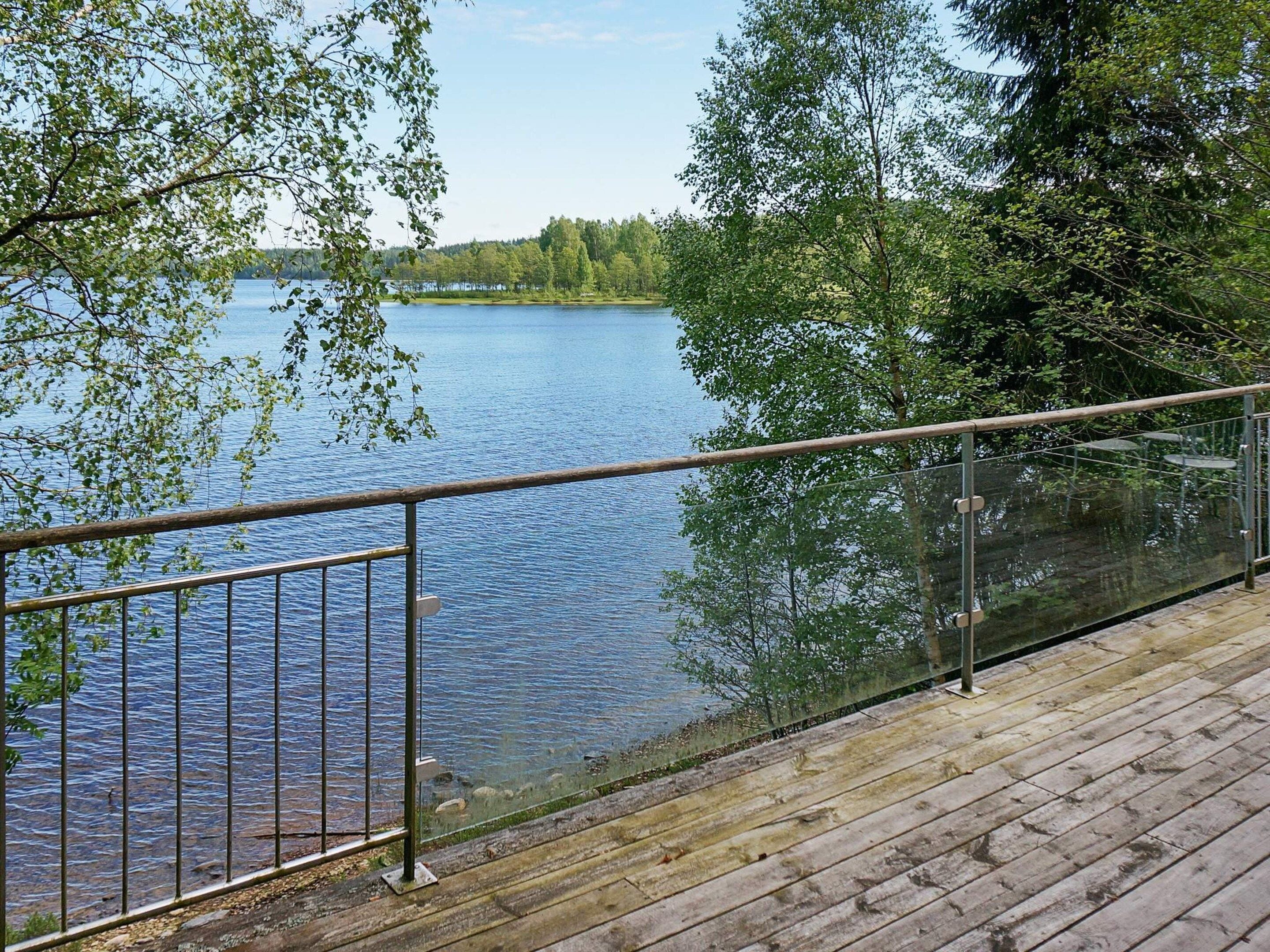 Une terrasse en bois avec une rambarde en métal, donnant sur un lac paisible, le tout entouré d’arbres.