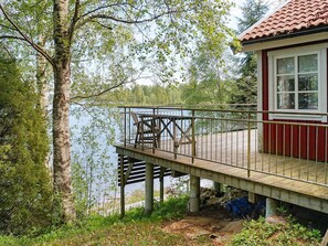 A wooden deck with a railing, a small building with a red exterior and white window frames, and a lake in the background.