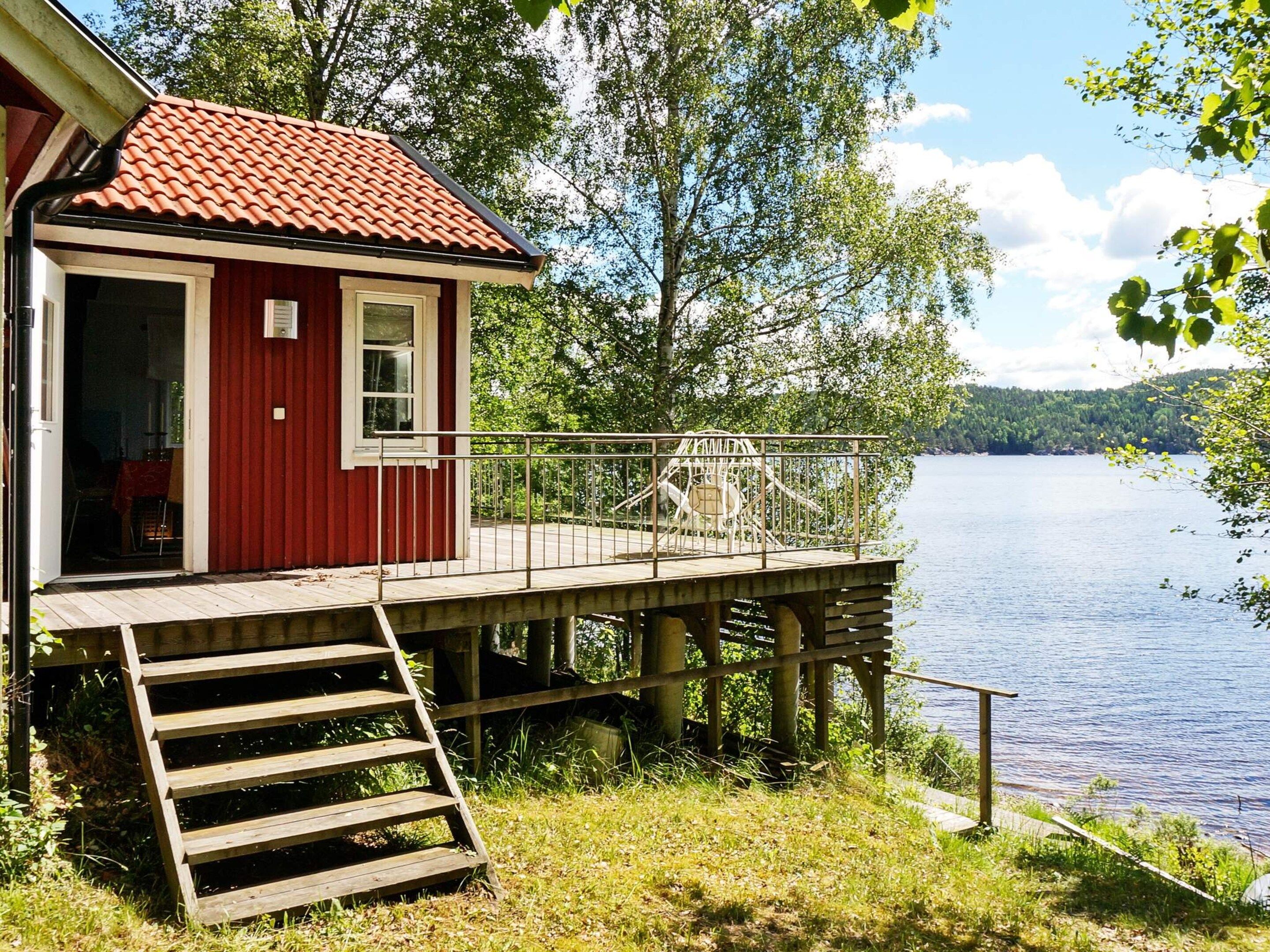 Une cabane rouge avec une terrasse en bois donnant sur un lac, une rambarde en métal et un escalier menant à l’entrée.