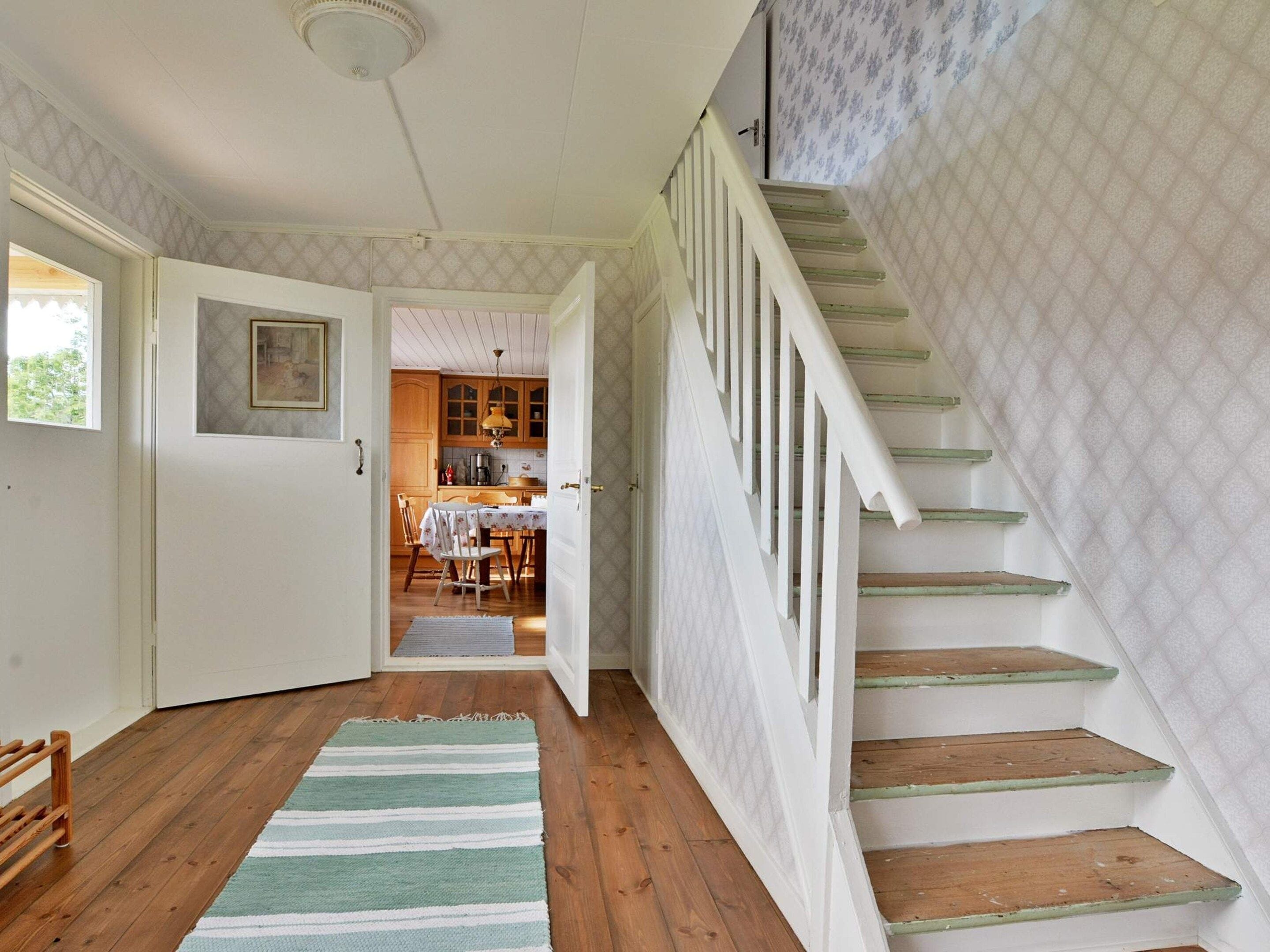 A staircase with white railings and wooden steps leading to a second floor. A wooden floor and a striped rug are visible in the foreground.