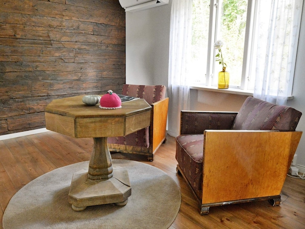 A room with a wooden floor, a round table with a vase and a crocheted item, a patterned sofa, and a window with curtains.