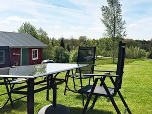 An outdoor dining area with a table and chairs, a grill, and a red building in the background. - Holiday Home in Håcksvik (Sandared)