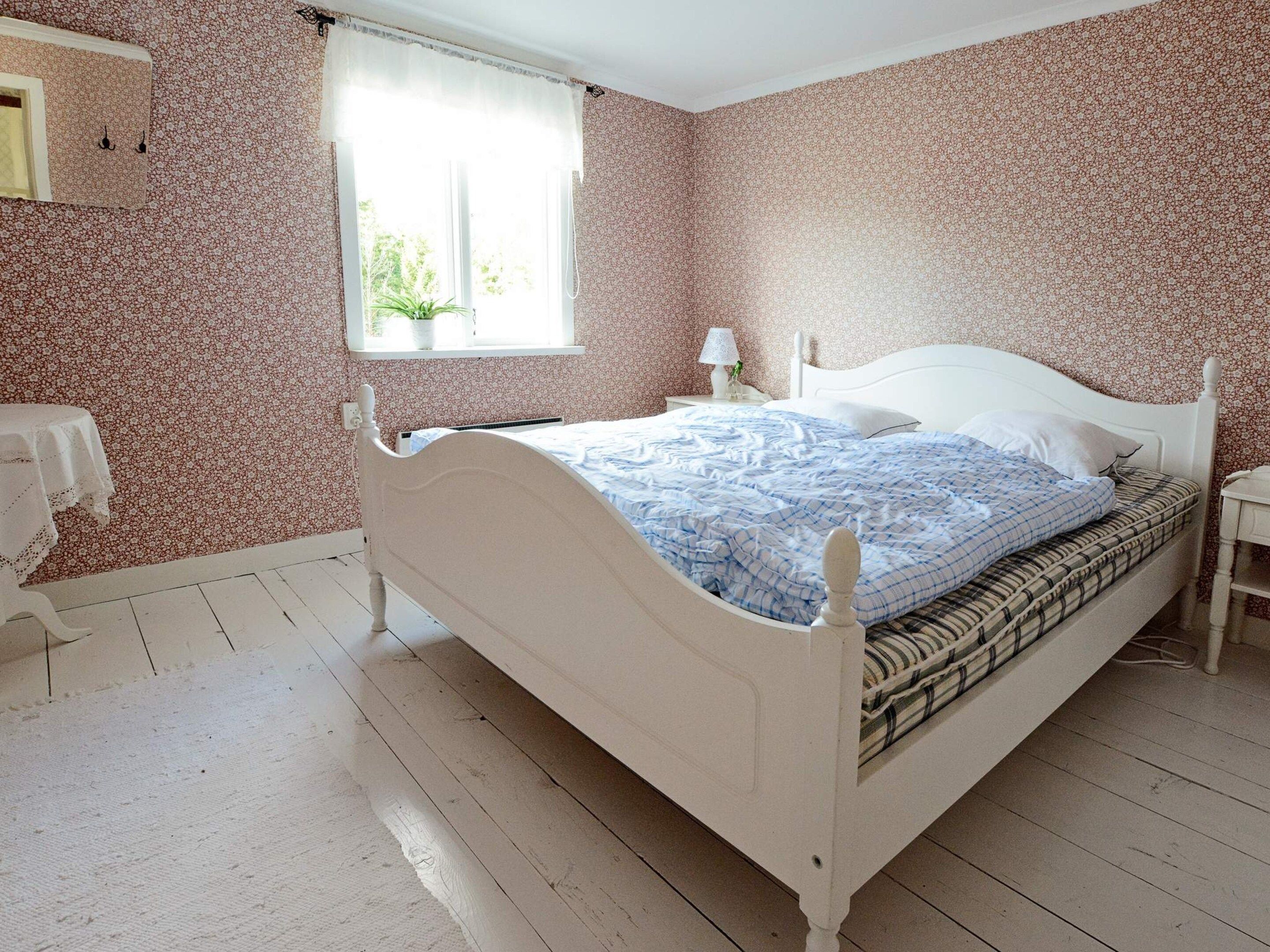 A bedroom with a white bed, a bedside table with a lamp, a window with a view of greenery, and a floral-patterned wall.