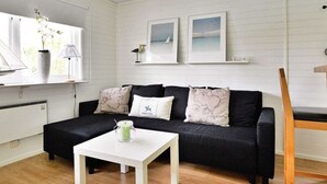 A modern living room with a black sofa, white coffee table, and a wooden chair. There are two framed pictures on the wall, a vase with flowers on the windowsill, and a lamp on the table. - Holiday Home in Gränna (Gränna)