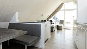 A modern loft-style room with a high ceiling, a dining area with a bench, a kitchenette, and a large window offering a view of the outdoors. - Holiday Home in Wendtorf (Wendtorf)