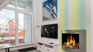 A modern living room with a fireplace, a flat-screen TV, and a large window overlooking a red building. - Holiday Home in Wendtorf (Wendtorf)