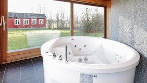 A modern bathroom with a large bathtub, wooden ceiling, and a view of a red house through large windows.