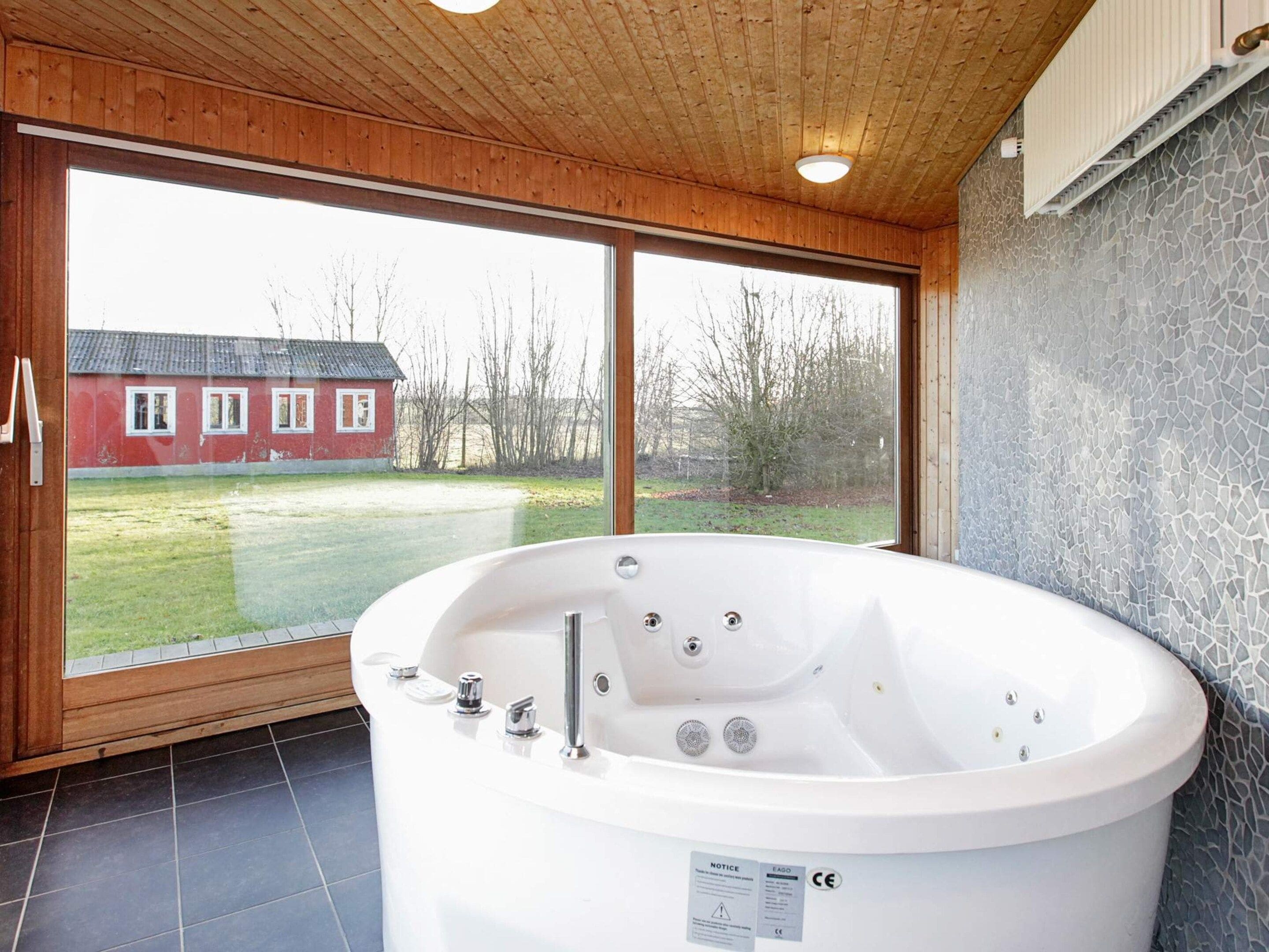 A modern bathroom with a large bathtub, wooden ceiling, and a view of a red house through large windows.