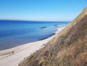A coastal landscape with a sandy beach, clear blue water, and a grassy cliff. - 5 Star Holiday Home in Frederiksvaerk (Frederiksvaerk)