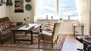 A cozy living room with a brown leather sofa, a wooden coffee table, a patterned rug, and a view of a snowy landscape through a large window with curtains. - 6 Person Holiday Home in Otta (Sel)
