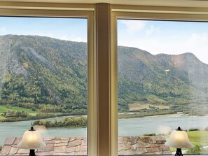A room with a large window overlooking a mountainous landscape with a river and a house in the distance. There are two lamps on the windowsill. - 6 Person Holiday Home in Otta (Sel)