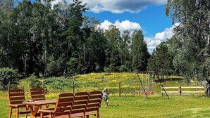 Une terrasse en bois avec une table et des chaises, entourée d’une pelouse agrémentée d’arbres et d’une balançoire.
